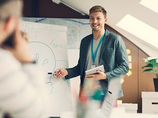 Ein junger Mann hält eine Präsentation vor einer Flipchart, auf der eine Glühbirne zu erkennen ist.