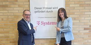 A man and a woman point to a sign that reads, "This lecture hall is funded by T-Systems."