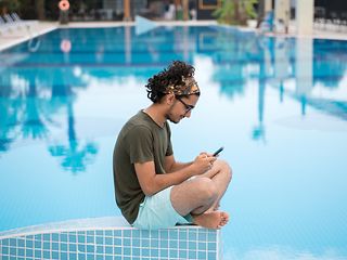 Mann mit einem Smartphone. Die Sonne scheint. Der junge Mann sitzt am Beckenrand eines Pools und surft mobil im Internet. 