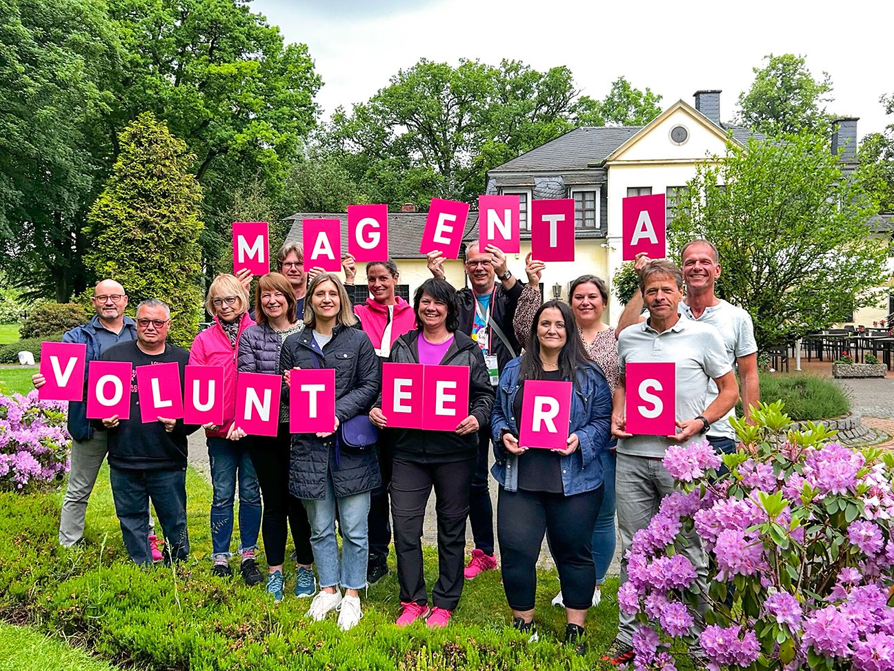 Telekom Mitarbeitende auf einem Grünfeld, halten die Buchstaben der Wörter „Magenta Volunteers“ in die Höhe.