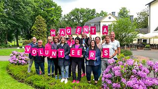 13 freiwillige Helfer und Helferinnen von Telekom halten ein Schild in der Hand, auf dem steht: Magenta Volunteers.