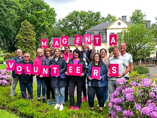 13 freiwillige Helfer und Helferinnen von Telekom halten ein Schild in der Hand, auf dem steht: Magenta Volunteers.