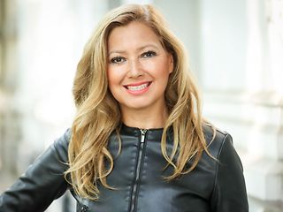 A woman with open blonde long hair and a black leather jacket smiles at the camera