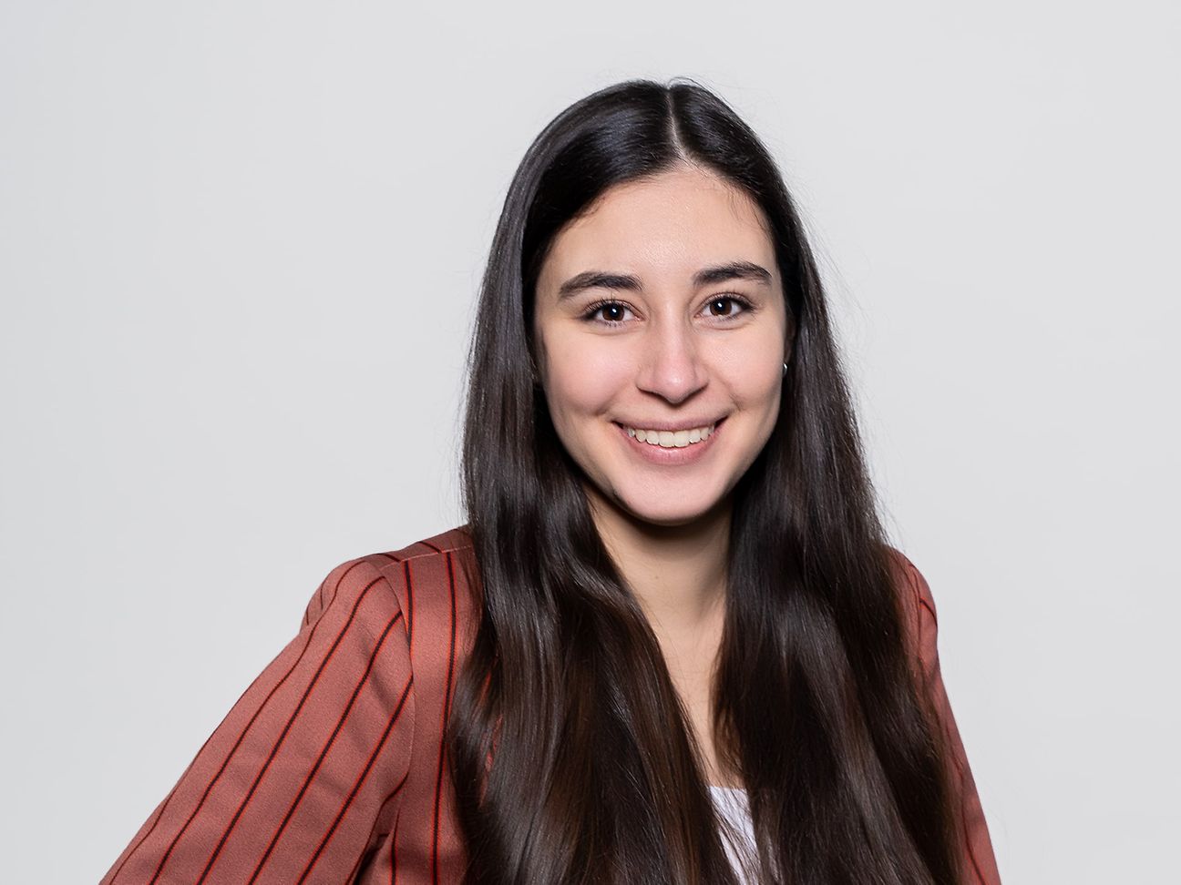 A young woman with long, black hair wears an orange-brown suit with black stripes and smiles into the camera.