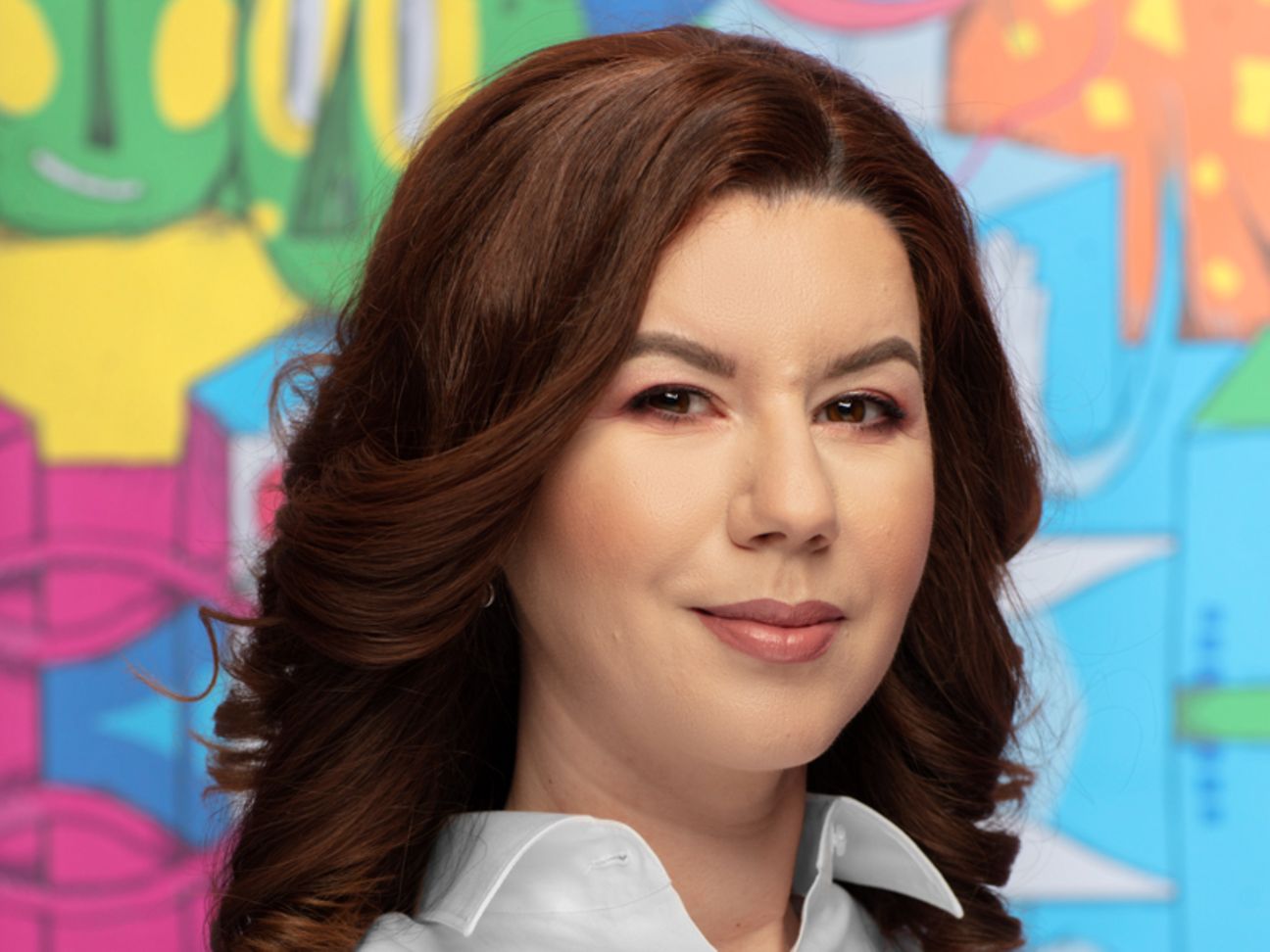 A young woman with brown wavy hair stands confidently in front of a colorful background and smiles slightly into the camera.