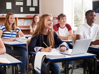 Jugendliche im Klassenzimmer schauen in Richtung Lehrkraft. Ein Mädchen hat einen Laptop vor sich auf dem Tisch.