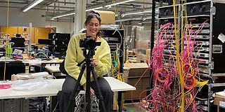 A woman sits behind the camera in a technical room