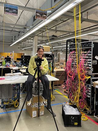 A woman sits behind the camera in a technical room