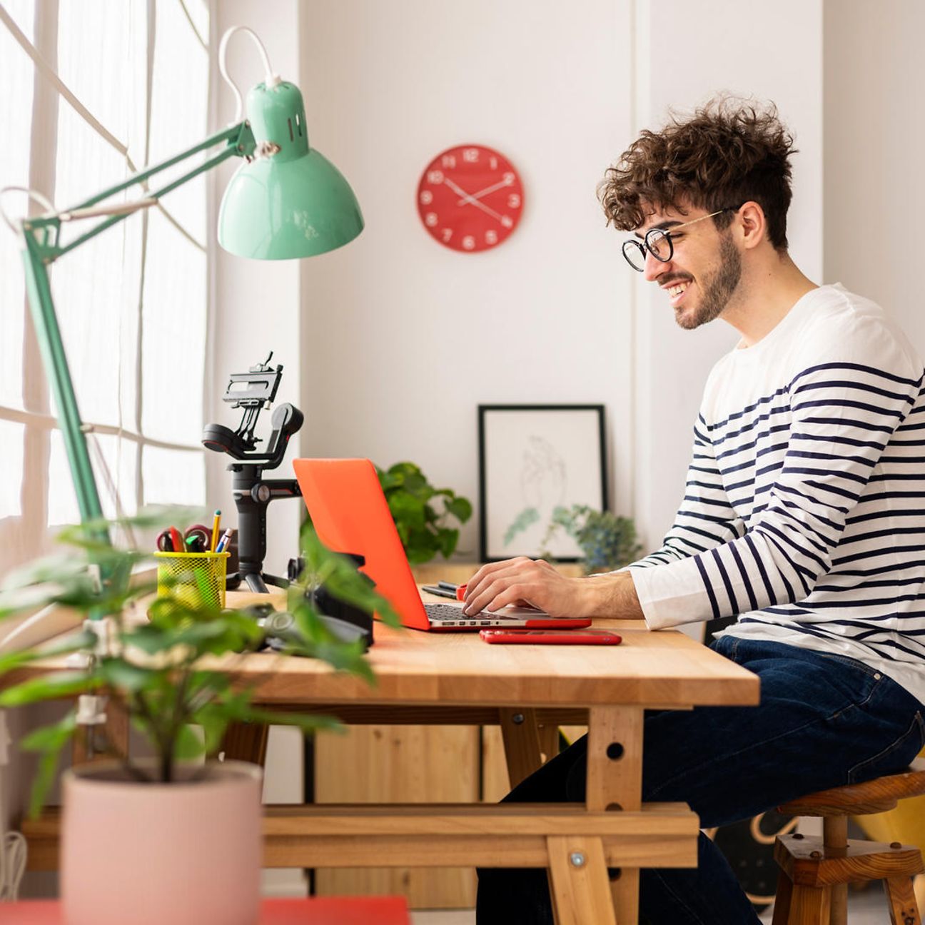 Ein junger Mann mit Brille und gestreiftem Pullover arbeitet lächelnd an einem Laptop in seinem Zuhause 
