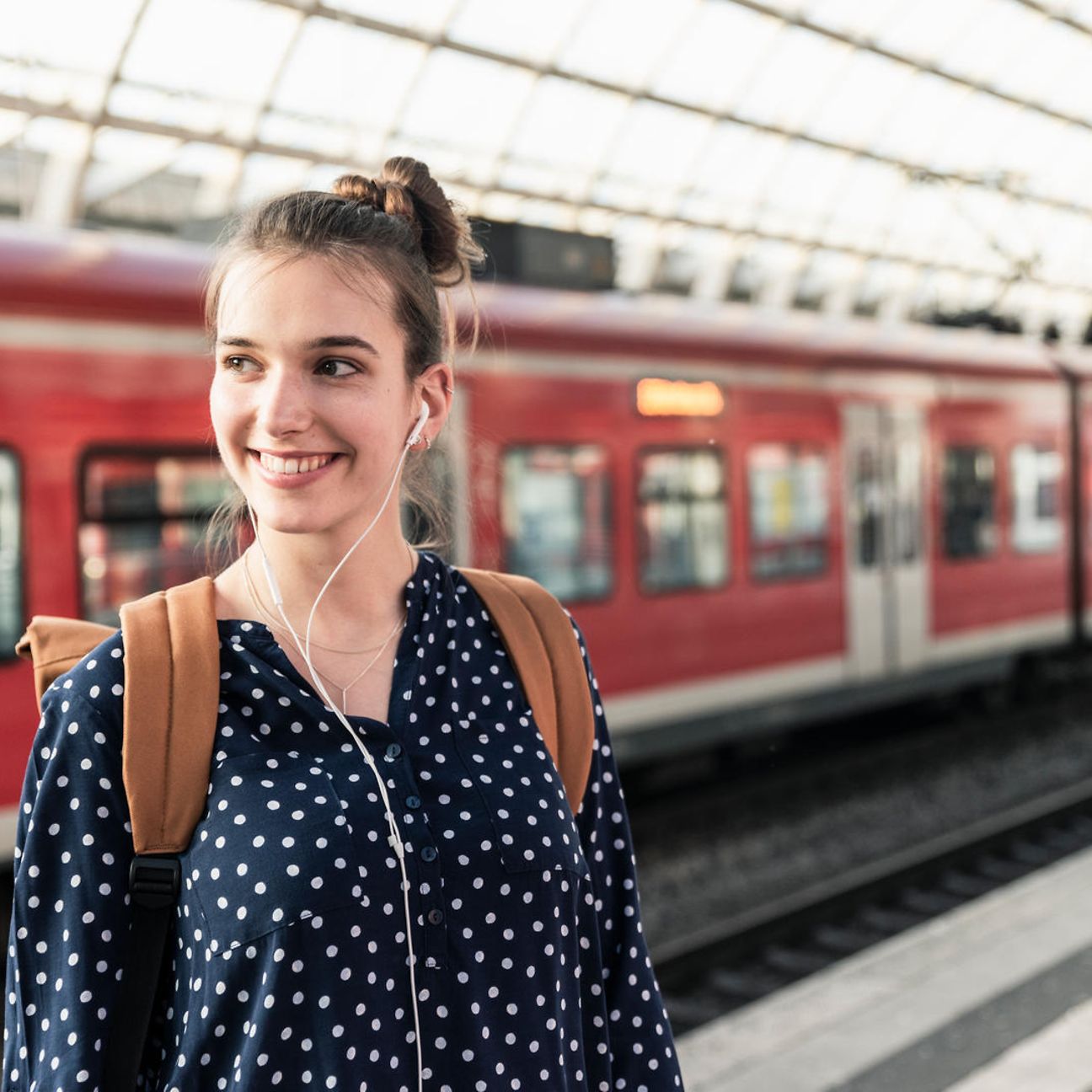 Eine junge Frau mit Rucksack und Kopfhörern steht am Gleis eines Bahnhofs.