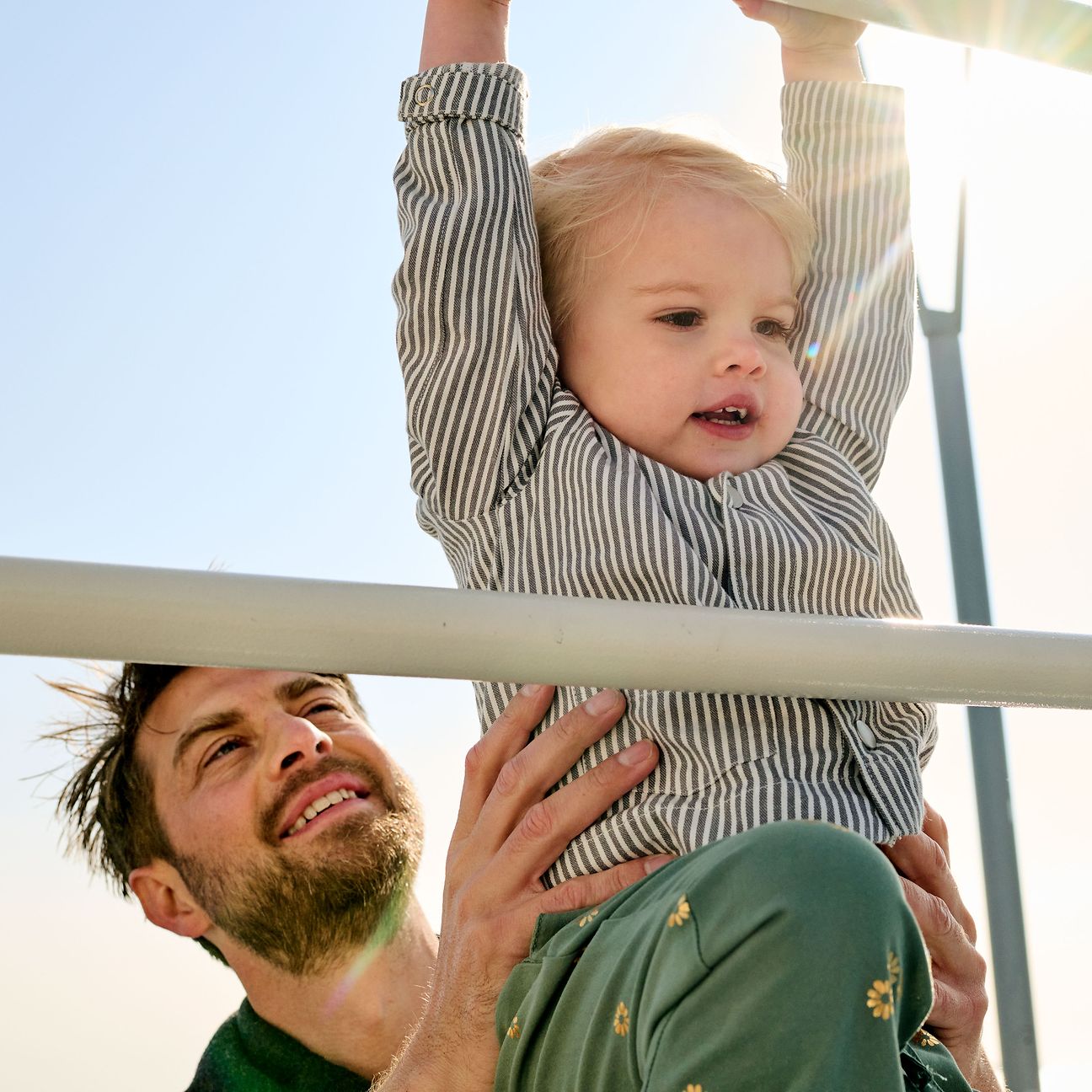 Ein Vater ist auf dem Spielplatz mit seinem Kleinkind und hebt es an ein Klettergerüst.