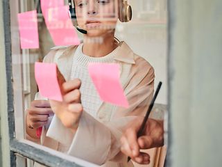 Eine Frau mit Kopfhörern klebt rosa Post-its an eine Scheibe, von rechts ist eine Hand mit einem Stift zu sehen