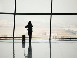 Eine Person mit Koffer steht an einem großen Fenster im Flughafen und blickt auf die Startbahn