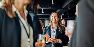 Young woman with champagne glass in hand stands in a group of people and laughs at other woman.