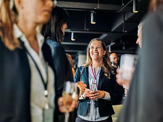 Young woman with champagne glass in hand stands in a group of people and laughs at other woman.