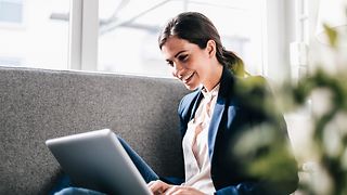 Eine gut gelaunte, junge Frau in Blazer sitzt auf einem grauen Sofa und sieht in ihren Laptop 