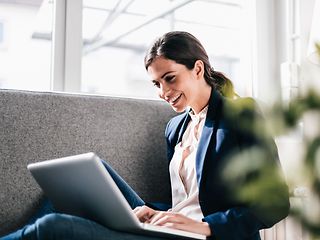 Eine gut gelaunte, junge Frau in Blazer sitzt auf einem grauen Sofa und sieht in ihren Laptop 