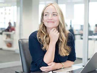 Junge Frau sitzt vor einem Laptop in einem Großraumbüro.