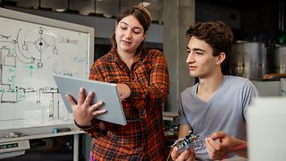 Two people are in a tech lab, holding a mini computer and a tablet