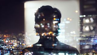 Shadowy depiction of a person reflected in front of a window, looking at a city in the dark