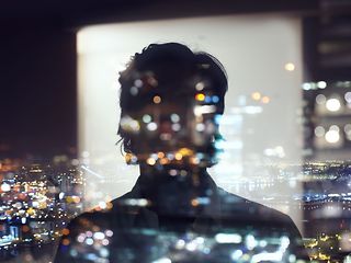 Shadowy depiction of a person reflected in front of a window, looking at a city in the dark