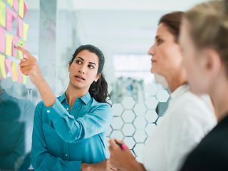 Gruppe von drei Frauen steht vor einer Pinnwand mit vielen Post-Its und diskutiert.