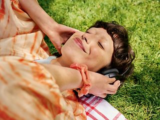 Junge Frau liegt mit Kopfhörern Musik hörend versonnen lächelnd im Gras.