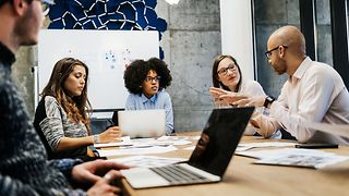 Several people are sitting at a table in a meeting room and are talking