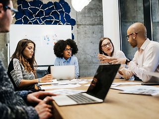 Several people are sitting at a table in a meeting room and are talking