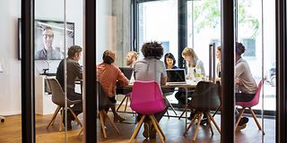 Group of people sitting at a table in a conference room working on laptops, a virtual participant can be seen on a large screen