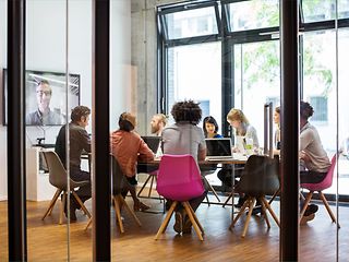 Group of people sitting at a table in a conference room working on laptops, a virtual participant can be seen on a large screen