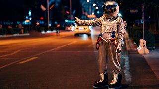 A hitchhiker in a space suit stands in the dark on a road and gives a thumbs up