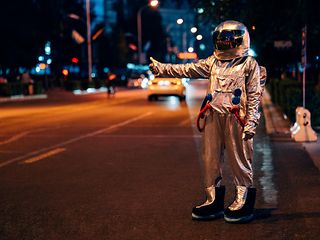 A hitchhiker in a space suit stands in the dark on a road and gives a thumbs up