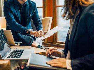 Two people sit opposite each other, while one of them gives the other something on a piece of paper