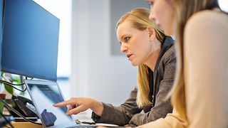 A woman explains something to another on a laptop in an office