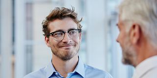 A young man in glasses smiles at an older man while they are talking in an office