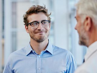 A young man in glasses smiles at an older man while they are talking in an office