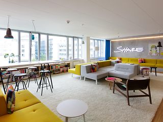 Modern, bright room with yellow sofas, high tables and chairs, and a neon sign stating “SHARE” on the wall