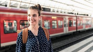 A young woman with a backpack and headphones stands on the platform of a train station.