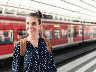 A young woman with a backpack and headphones stands on the platform of a train station.