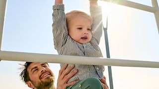 A father is at the playground with his toddler and lifts him onto a climbing frame.