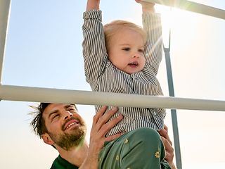 A father is at the playground with his toddler and lifts him onto a climbing frame.