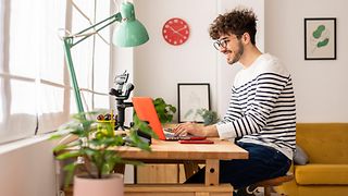 A young man with glasses and a striped sweater works smiling on a laptop in his home