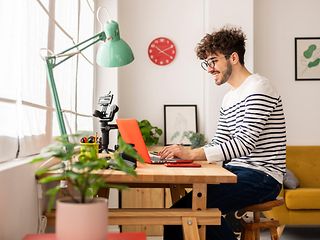 A young man with glasses and a striped sweater works smiling on a laptop in his home