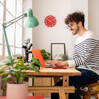 A young man with glasses and a striped sweater works smiling on a laptop in his home