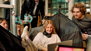 A young woman sits smiling on a beanbag and looks at her laptop, a young man stands next to her.