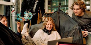 A young woman sits smiling on a beanbag and looks at her laptop, a young man stands next to her.