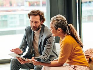 A woman and a man sitting in a lounge area talking while looking at a tablet