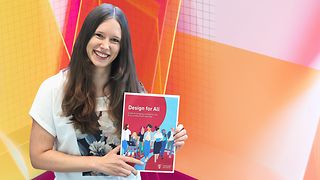 Annika Theiß hält den „Design für Alle Guide“ in der Hand. Sie steht vor einem bunten Design-Hintergrund.