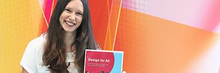 Annika Theiß hält den „Design für Alle Guide“ in der Hand. Sie steht vor einem bunten Design-Hintergrund.